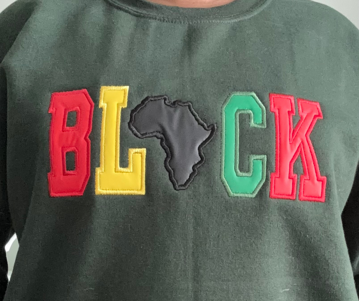 Dark green crewneck sweatshirt featuring bold, varsity-style appliqué lettering that spells out “BLACK.” The letters are in red, yellow, and green, with the letter “A” replaced by a black embroidered shaped of the African continent.