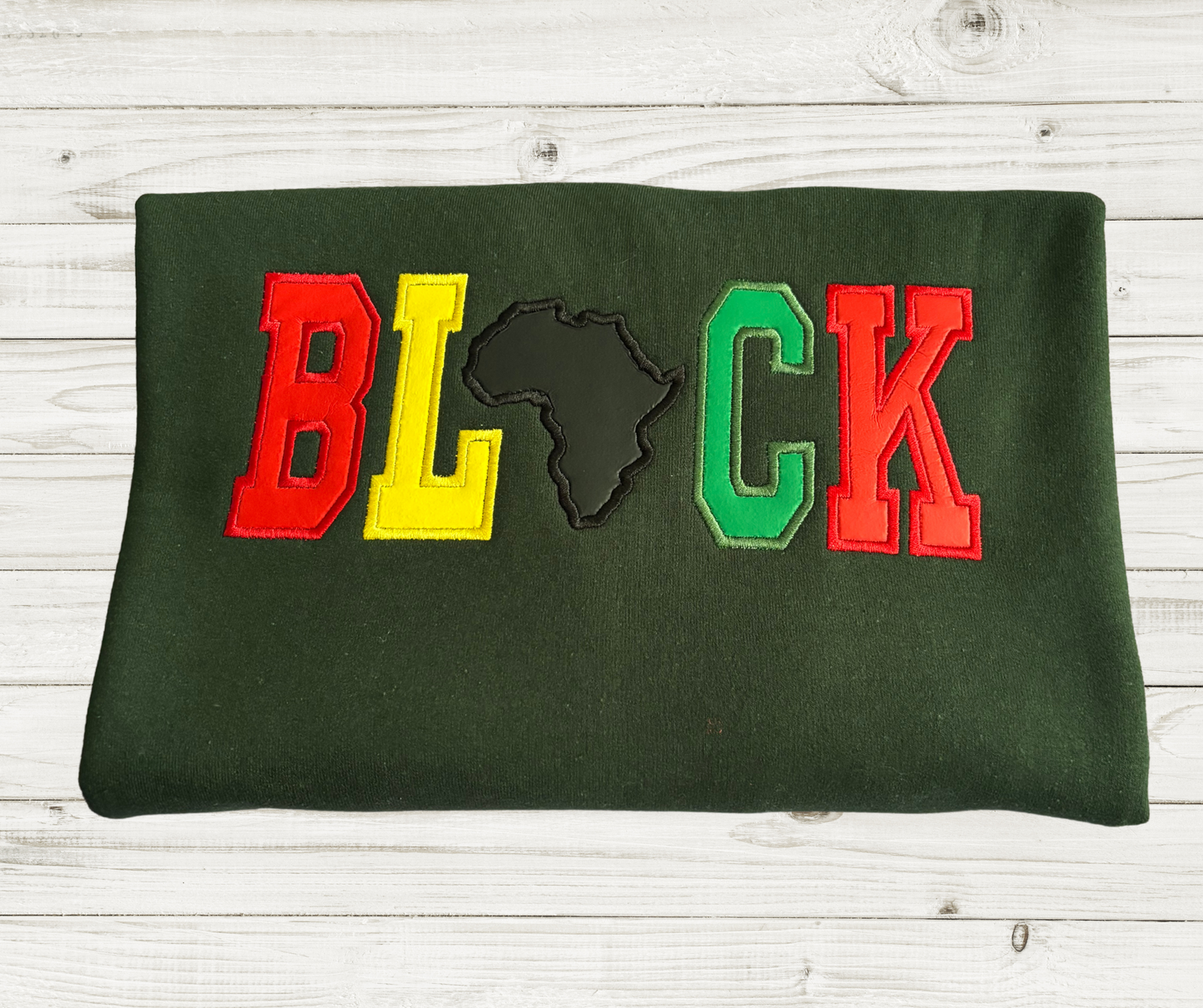 Dark green crewneck sweatshirt featuring bold, varsity-style appliqué lettering that spells out “BLACK.” The letters are in red, yellow, and green, with the letter “A” replaced by a black embroidered shaped of the African continent.